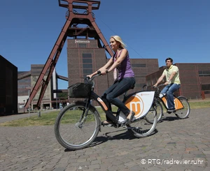 Ein Paar ist unterwegs auf Rädern von Metropolrad, im Hintergrund sieht man die Zeche Zollverein.